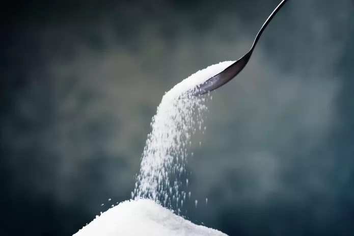 A spoon pours granulated sugar onto a pile, against a dark blurred background.