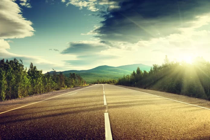 A scenic view of an open road leading towards mountains under a sunny sky with lush greenery on both sides.