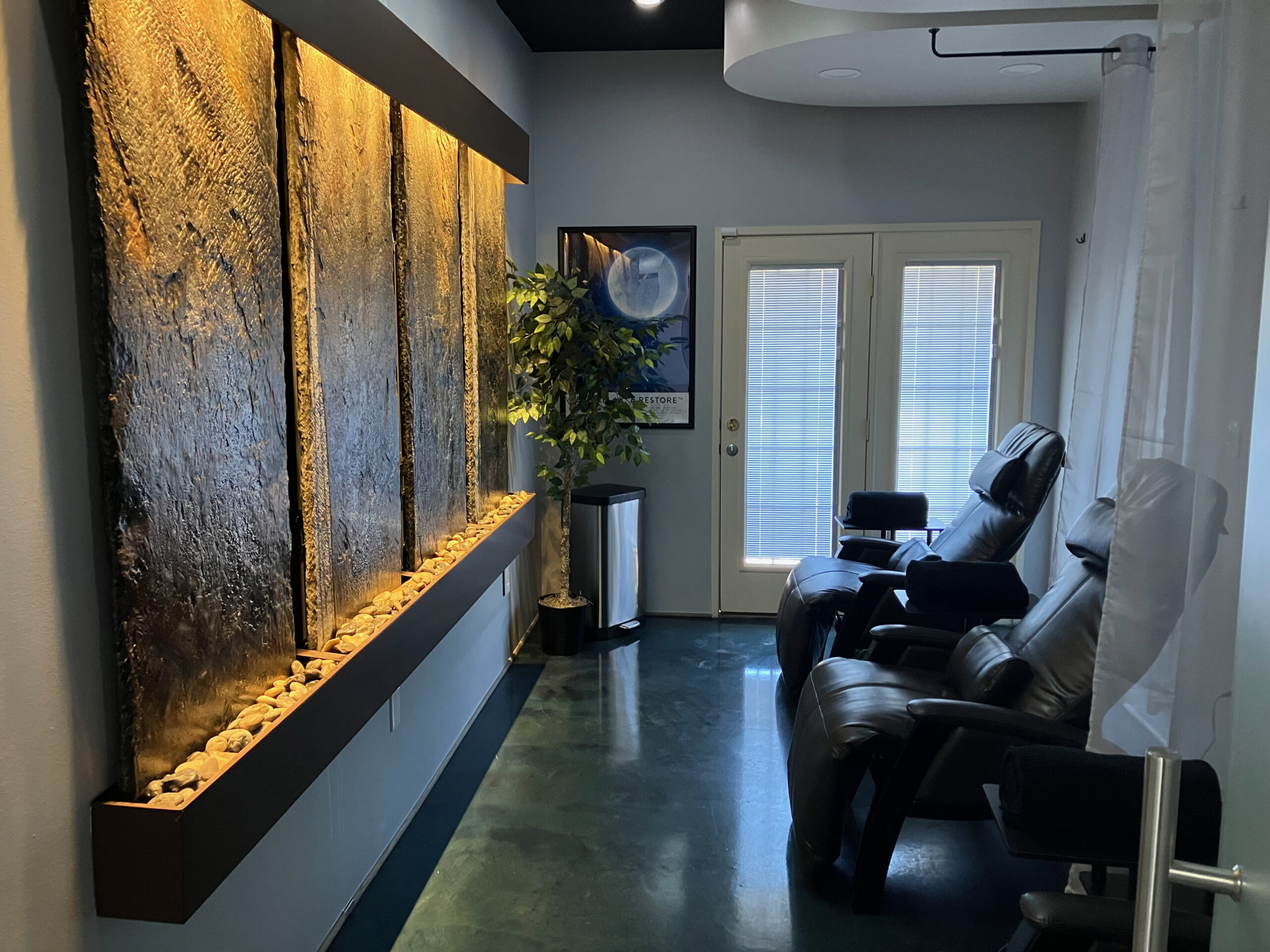 A tranquil room with leather recliner chairs faces a textured wall installation with built-in lighting and stones. A potted plant near the closed double glass doors adds a touch of nature, creating an inviting atmosphere perfect for IV Nutrition El Paso sessions.