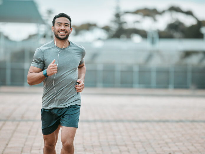 A man jogging outdoors in a grey shirt and shorts, wearing earphones and smiling.