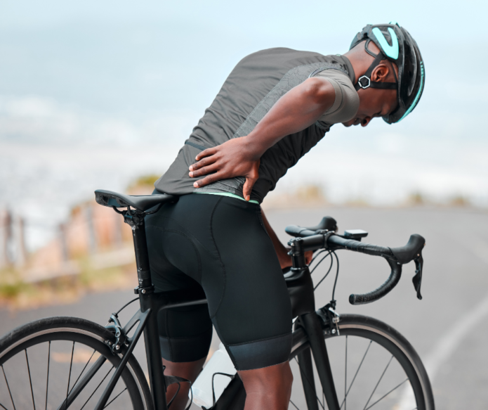 A person in cycling gear stands beside a bicycle, holding their lower back with one hand and looking downward.