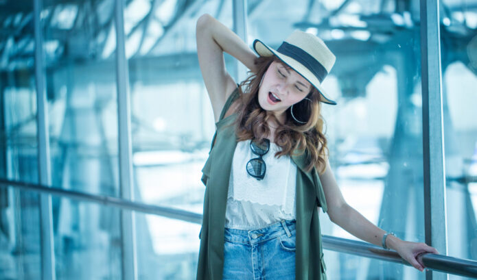 A woman in a hat and casual clothes stands near a glass wall, stretching and yawning with one hand behind her head and sunglasses hanging from her top.