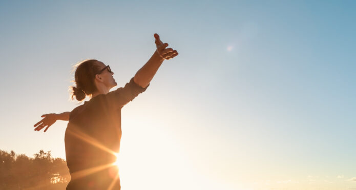 A person with sunglasses is standing with arms outstretched, facing the sunset or sunrise with a clear blue sky in the background.