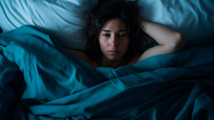 A person lies in bed with teal-colored sheets and white pillows, looking directly at the camera with a neutral expression. Their arms are partially visible, resting on the pillow and bed.