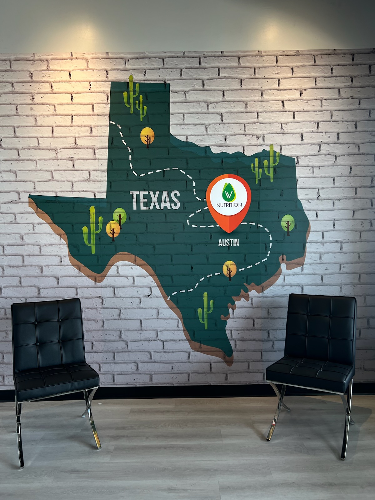 A wall mural of Texas with "Austin" marked, featuring a path and cacti, creates a serene backdrop behind two black chairs—perfect for a relaxing session of IV Drip Therapy in Austin.