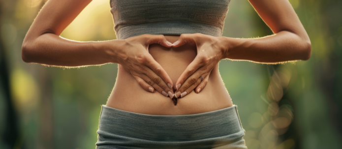 A person wearing a grey sportswear forms a heart shape with their hands over their bare midriff against a blurred outdoor background.