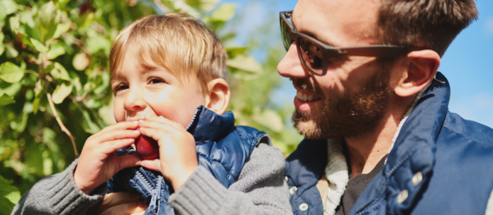 A man wearing sunglasses holds a young child who is eating an apple. They are both dressed warmly and standing outdoors on a sunny day.