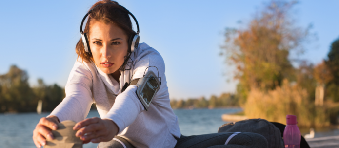 Woman wearing headphones and sportswear stretches while sitting outdoors near a body of water. A water bottle and bag are nearby.