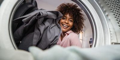 A woman with curly hair is smiling while putting laundry into a washing machine, viewed from inside the machine.