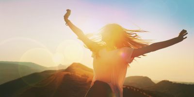 A person stands with arms outstretched, backlit by the sun, against a backdrop of mountains and clear sky.