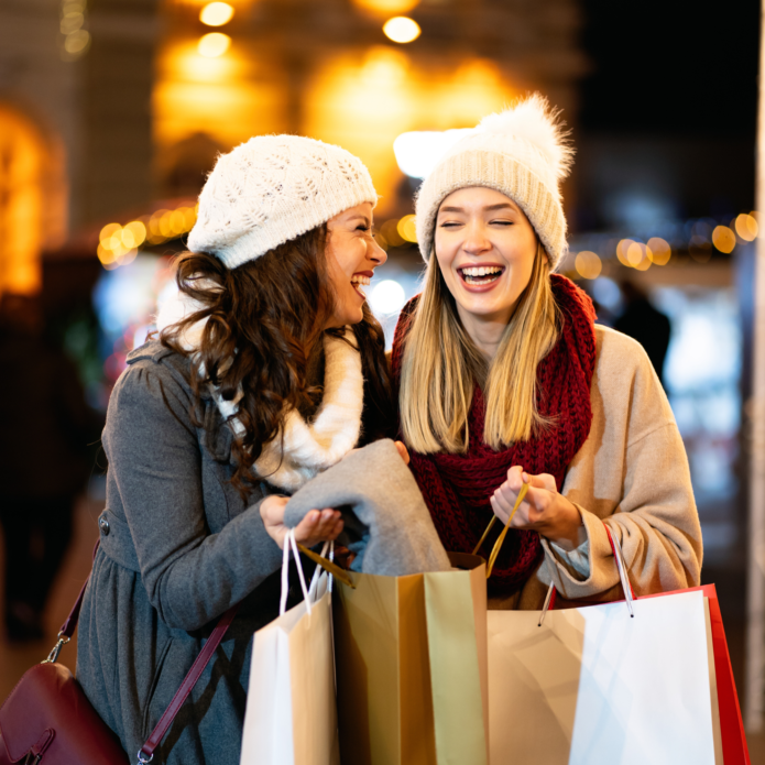 Two women in winter clothing, energized by nutrient therapy, laugh and hold shopping bags outdoors at night.