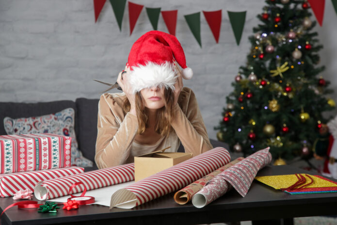 Person in a Santa hat looks tired while sitting at a table cluttered with wrapping paper and gift boxes. A decorated Christmas tree and festive banners are in the background.