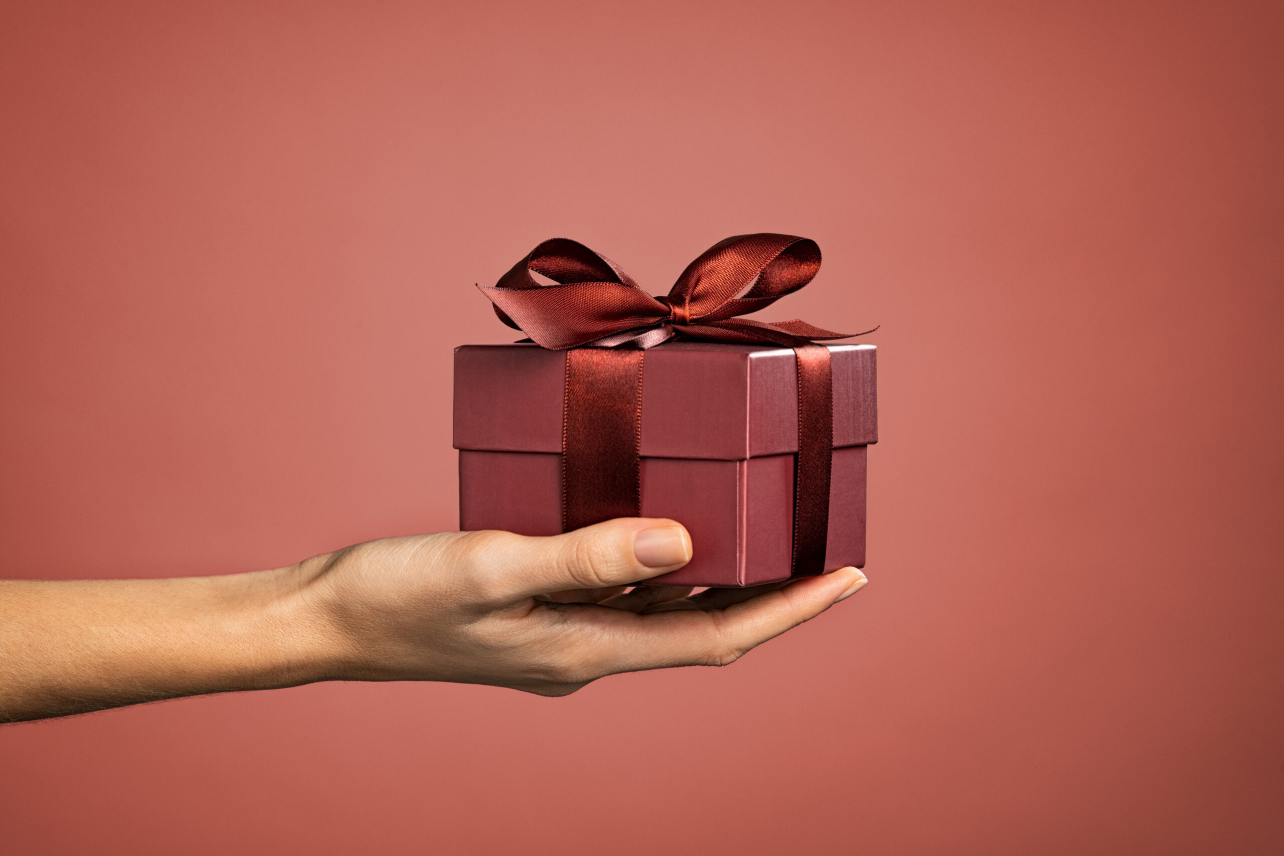 A hand holds a small, closed gift box with a shiny maroon ribbon on a dark peach background.