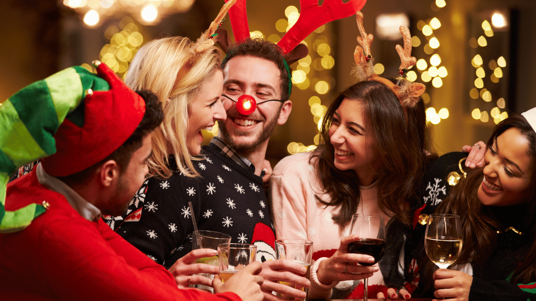 Five people wearing festive attire and reindeer antlers laugh together while holding drinks in a warmly lit setting.