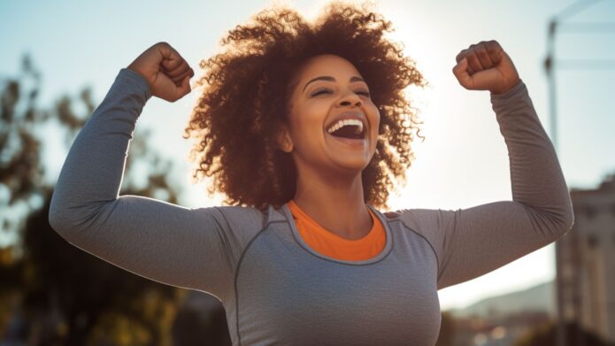 Person outdoors with curly hair, smiling and raising their arms in a triumphant pose. Wearing a gray long-sleeve shirt and orange top, with sunlight in the background.